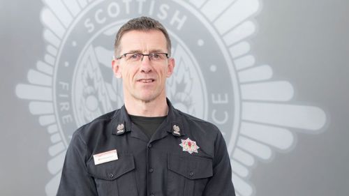 Deputy Assistant Chief Officer Stephen Wood in uniform in front of a grey SFRS crest