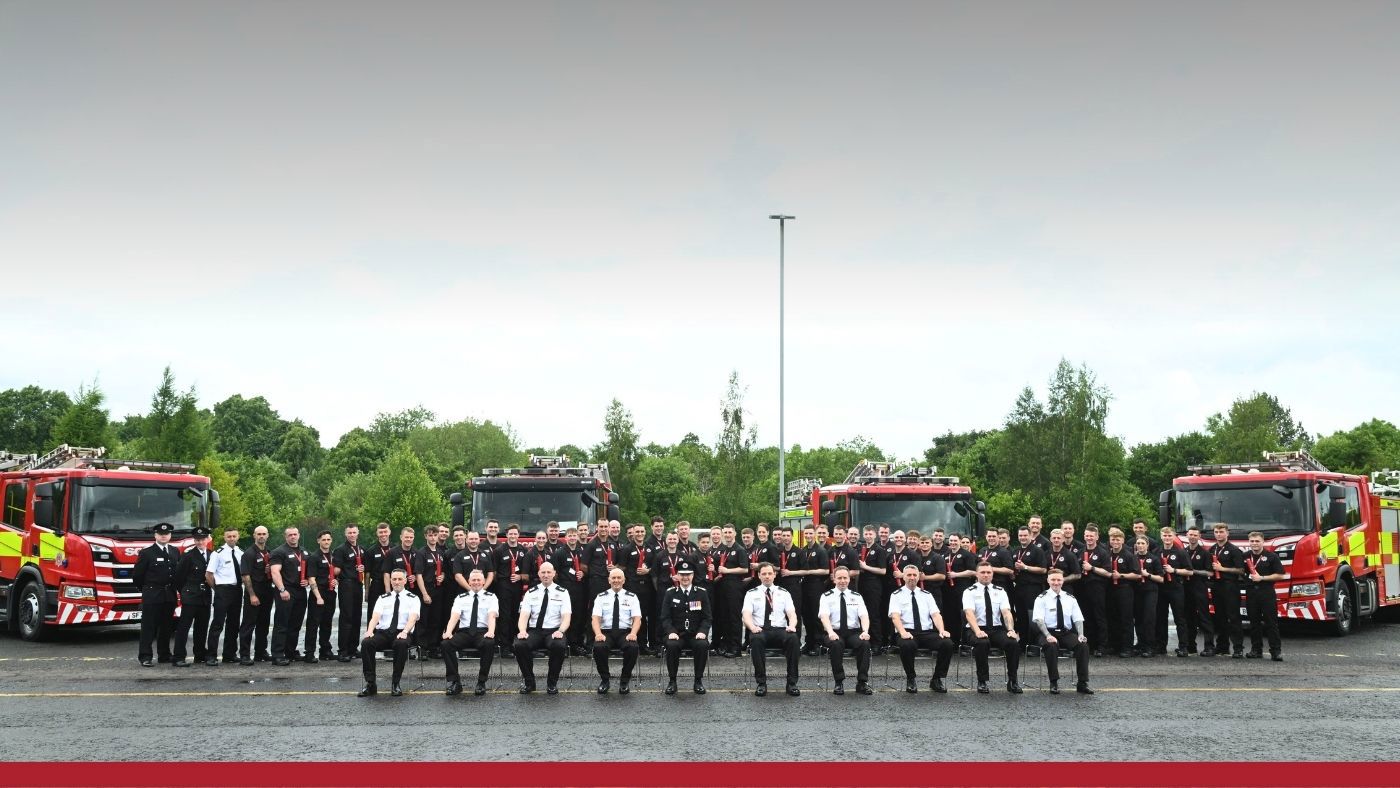 A group of firefighters standing in front of four fire trucks, dressed in a black uniform, smiling and posing for the photo.