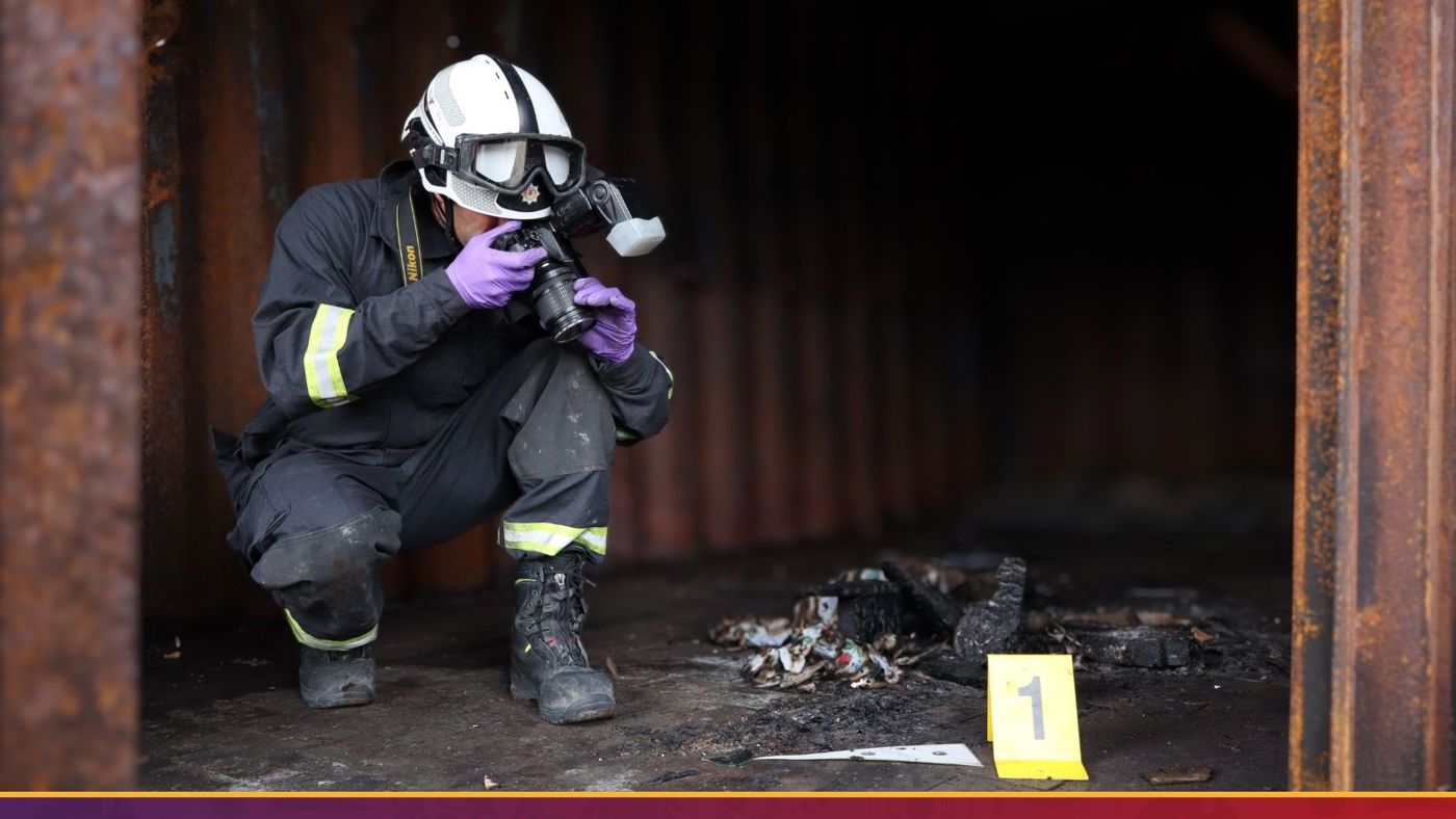 A firefighter crouches beside a pile of debris, surveying the area with a focused expression.