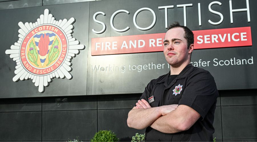 Firefighter standing confidently with arms crossed in front of Scottish Fire and Rescue Service sign. He wears a black uniform, conveying dedication and pride.