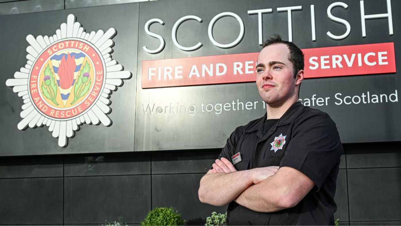 Firefighter standing confidently with arms crossed in front of Scottish Fire and Rescue Service sign. He wears a black uniform, conveying dedication and pride.