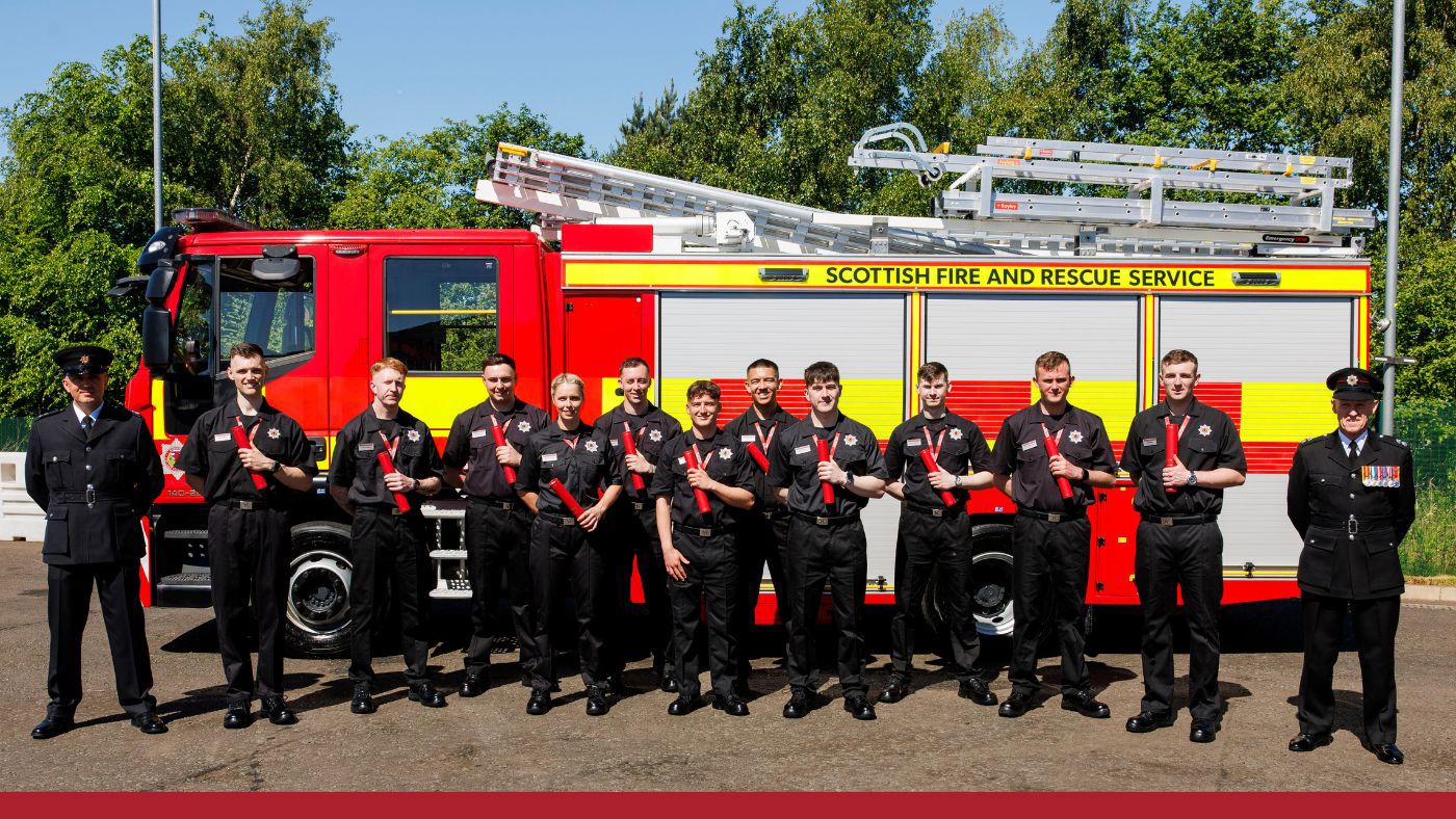 A team of firefighters dressed in black uniforms stands alongside a fire truck.