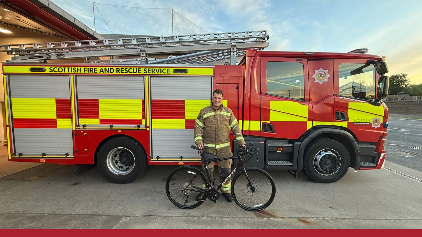 A man in a firefighting uniform stands beside a bicycle, smiling and posing for the camera.