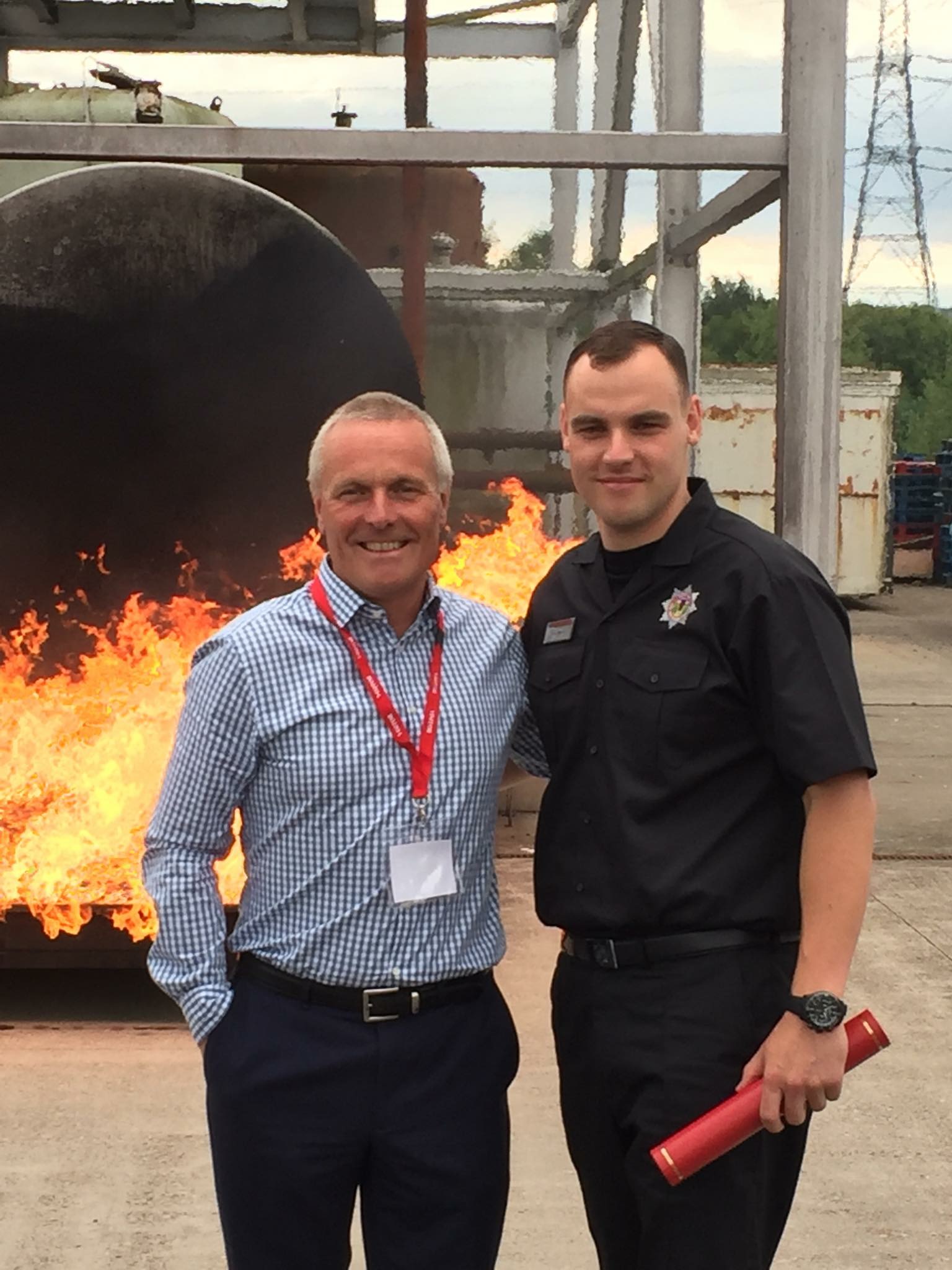 Picture of a man in a checkered shirt standing with a firefighter in uniform with a fire behind them