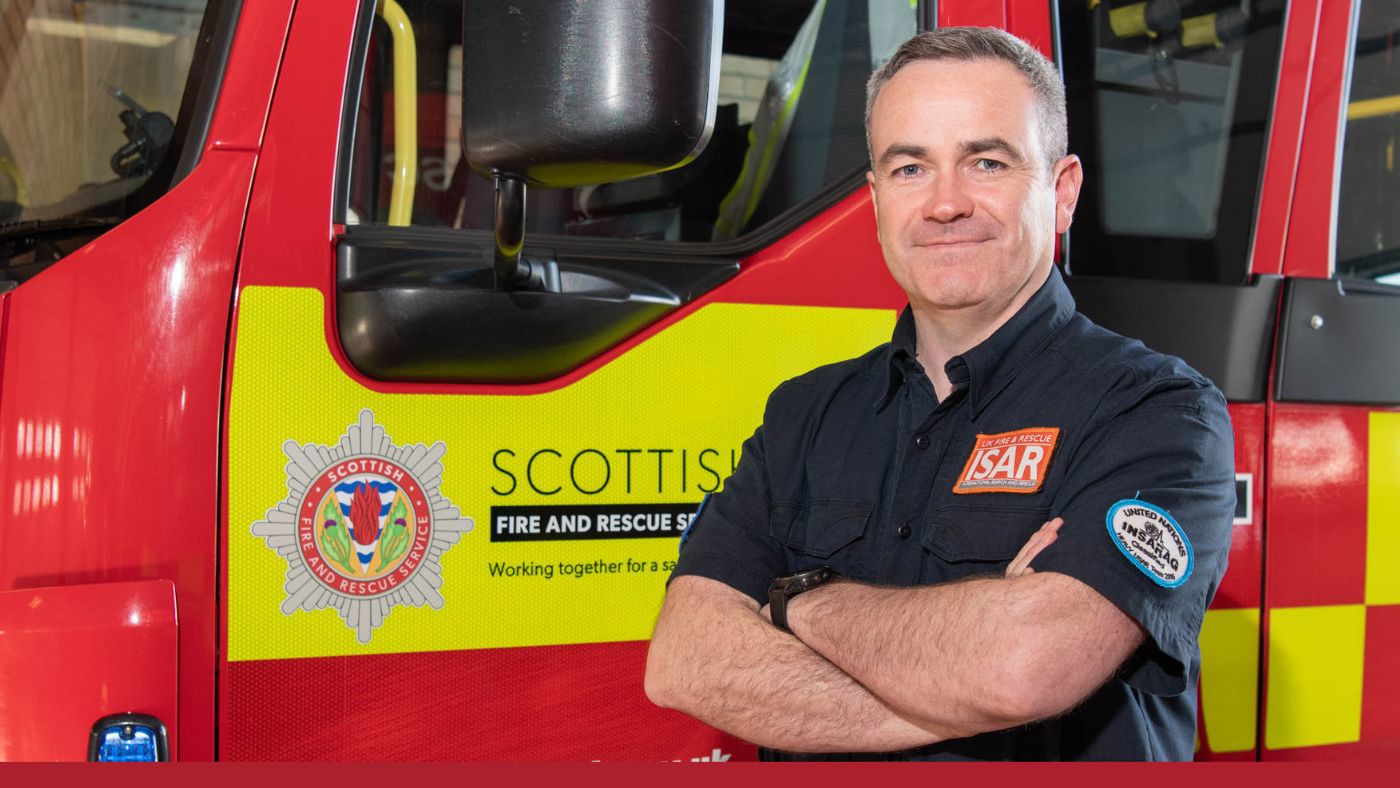 A firefighter in uniform stands beside a red fire appliance.