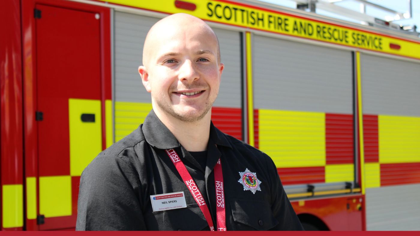 A man in a black shirt and red tie stands confidently in front of a fire appliance.
