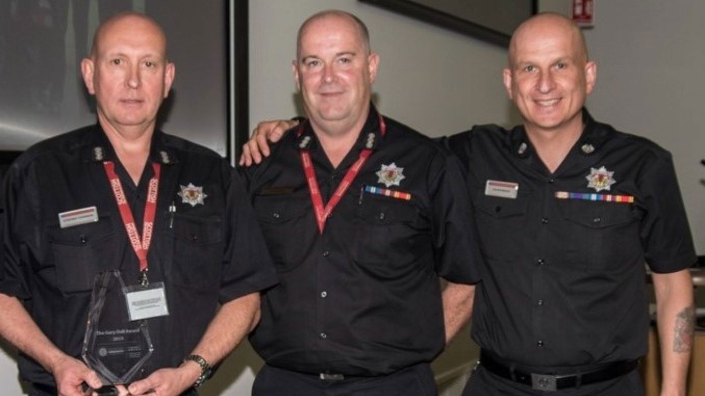 Three firefighters in black uniform posing with a trophy in hand.