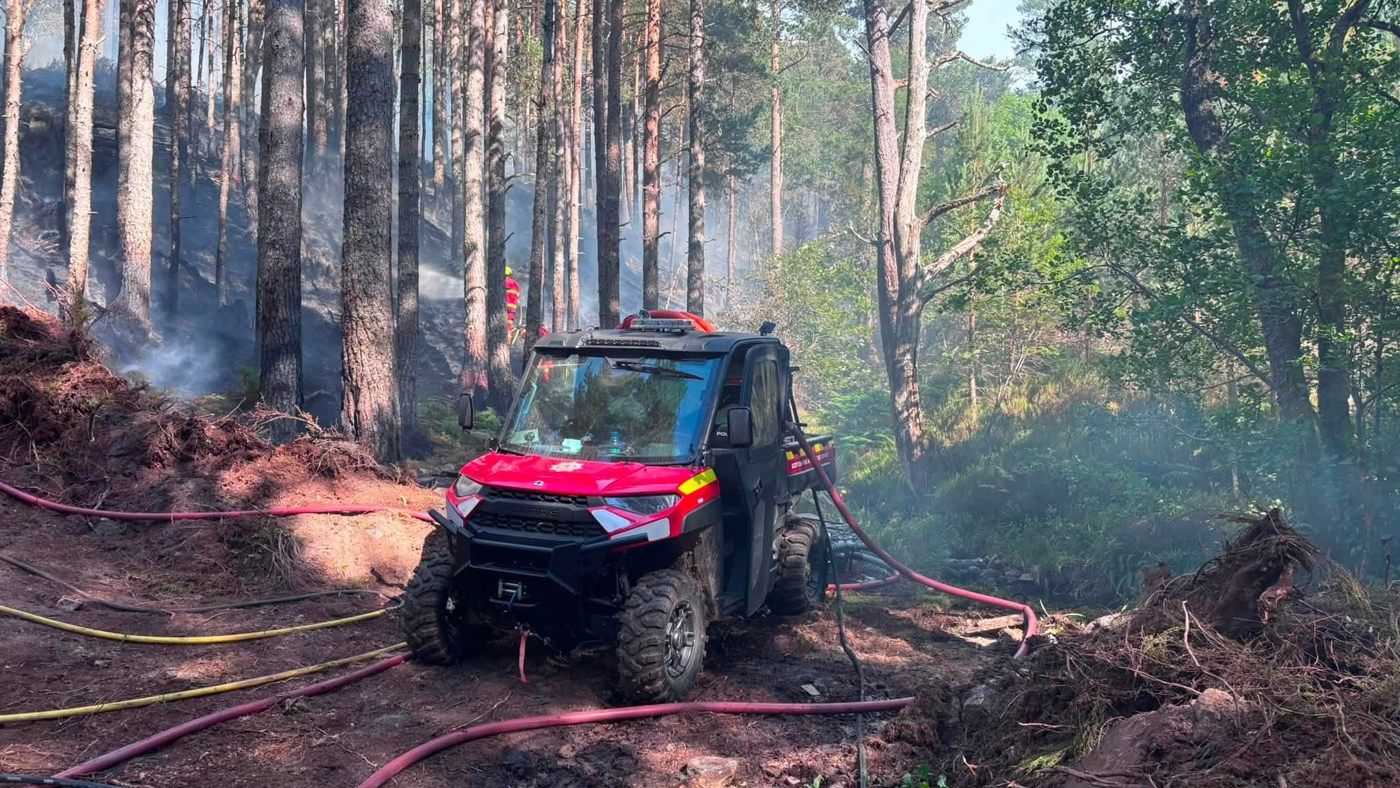 A small red all terrain vehicle with thick tyres and multiple hoses running across the ground in a woodland setting assiting firefighters with a wildfire 