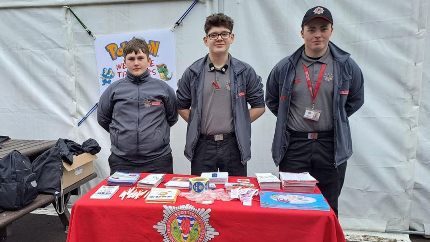 Three people in uniform stand behind a table with promotional items under a tent. A sign with "Pokémon" is in the background.