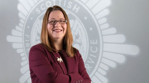 Head and shoulders image of Chair of the Board, Dr Kirsty Darwent smiling in front of a grey SFRS crest