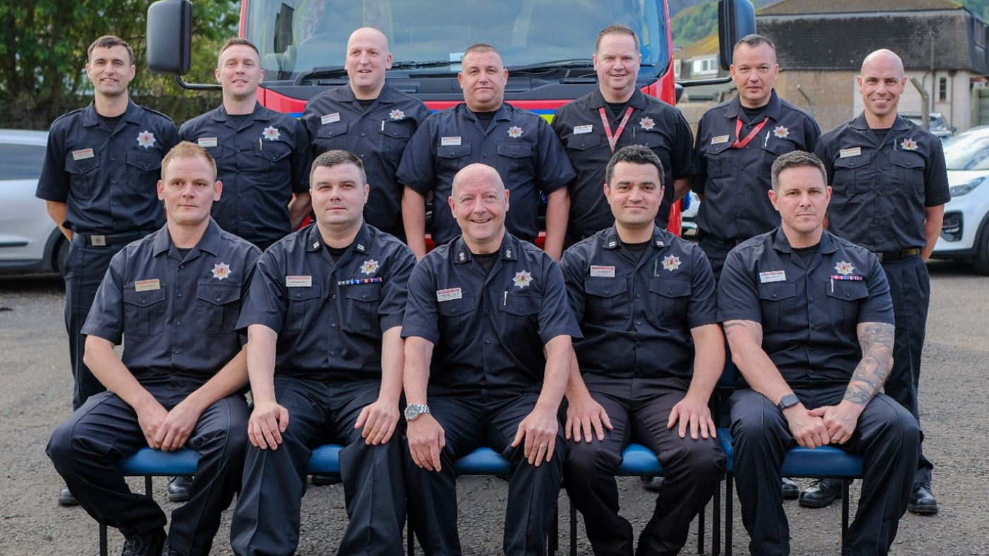 Group photo including 12 members of the crew from Tillicoultry Community Fire Station, Colin Hutcheson is sitting in the centre, with a fire appliance in the background celebrating Colin Hutcheson's 40 years of service
