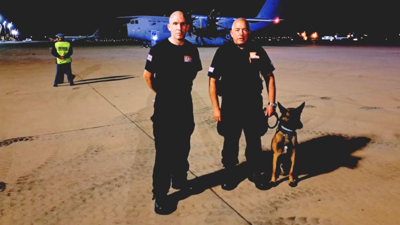 two firefighters wearing black uniforms standing on a airport runway at night with a service dog