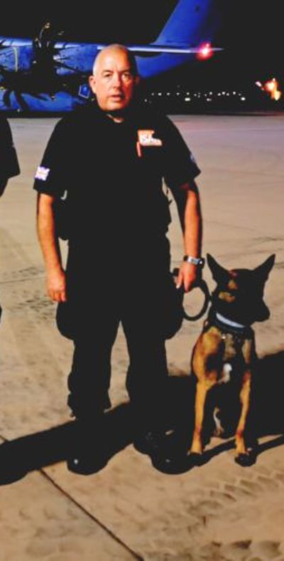 two firefighters wearing black uniforms standing on a airport runway at night with a service dog