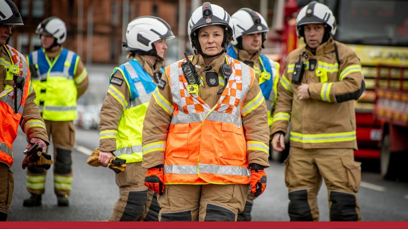 A group of firefighters in full gear and helmets on a street. A woman firefighter in an orange vest stands in front, appearing focused. A fire appliance is in the background.