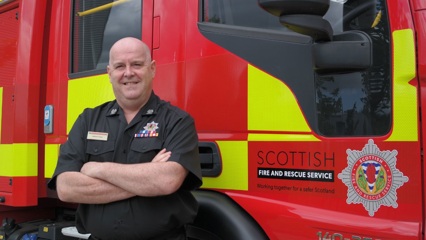 A fire officer in a black uniform standing next to a red fire appliance.