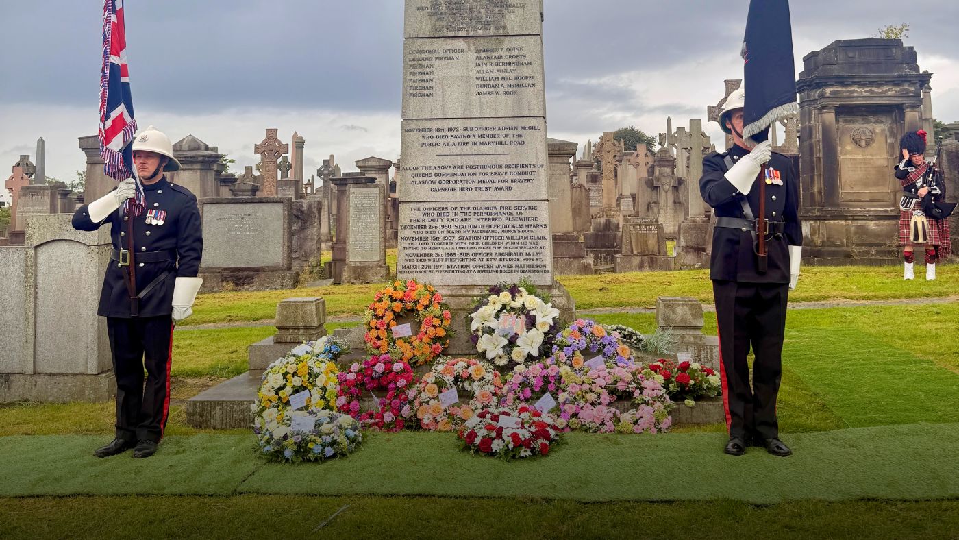 two firefighters in ceremonial dress stand by a gravestone covered in wreaths 