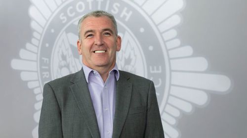 Mark McAteer smiling in a grey suit standing front of a grey SFRS crest