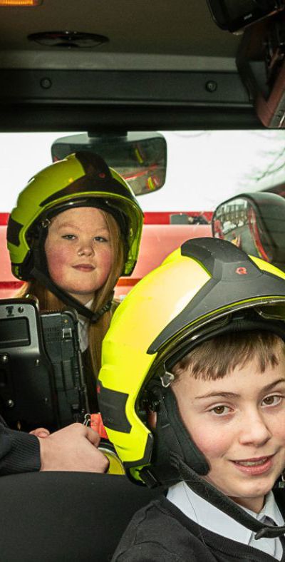 Three children sit in front of fire appliance with firefighter helmet on.