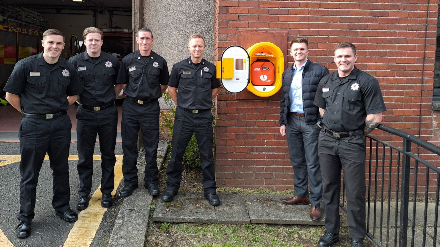 Six men, five in black uniforms and one in a suit, standing next to a newly installed defibrillator on a brick wall outside a fire station building.