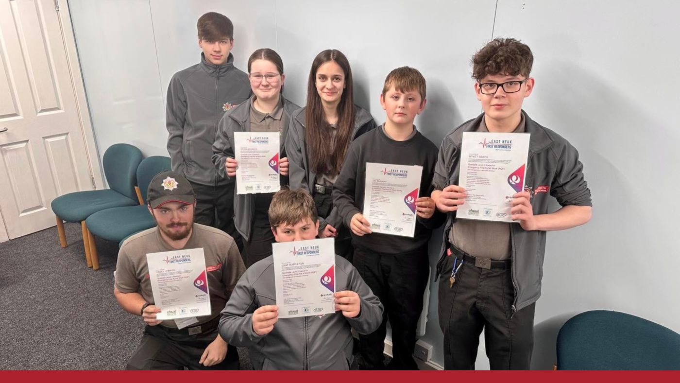 Seven young people in matching grey uniforms pose with certificates in a room with green chairs.