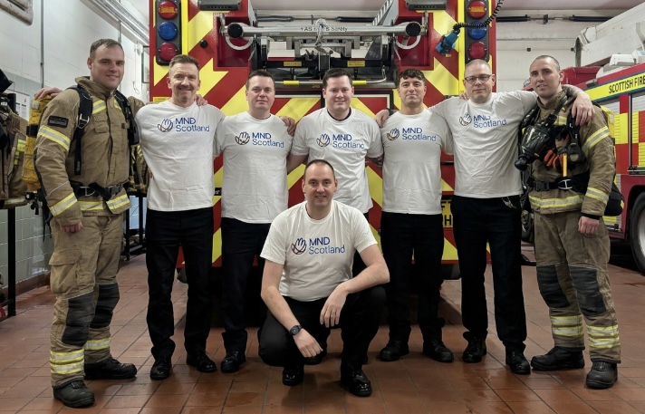 Group of firefighters in fire kit and MND Scotland t-shirts standing in front of fire appliance.