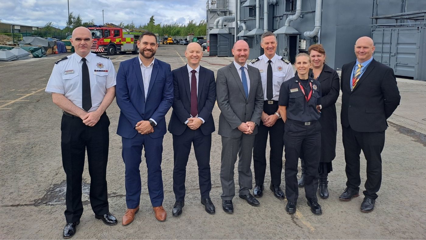 Group photo of people standing in a row smiling wearing smart clothing with the new Perth training facility and a fire appliance in the background