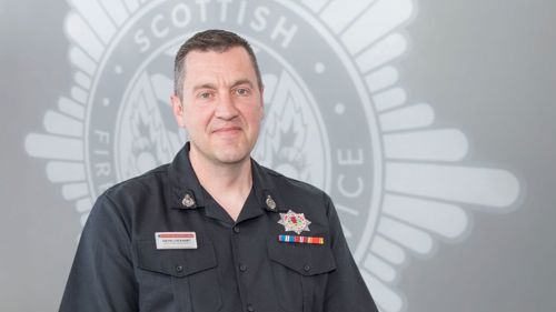 David Lockhart in black SFRS uniform standing in front of a grey SFRS crest