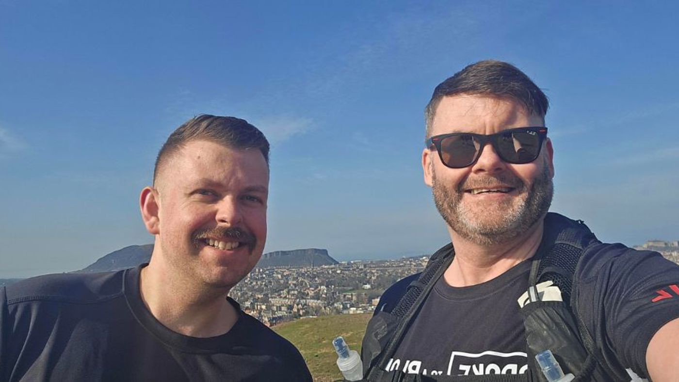 Two men smiling and taking a selfie together on a scenic hilltop with a clear sky in the background.